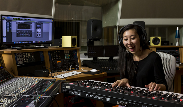 A woman in a black shirt with headphones on is playing the keyboard while sitting in a recording studio